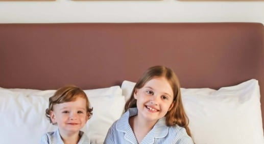 Two children in striped pajamas sit on a bed with a fluffy gray dog, smiling in a bright bedroom with abstract framed art above the headboard.