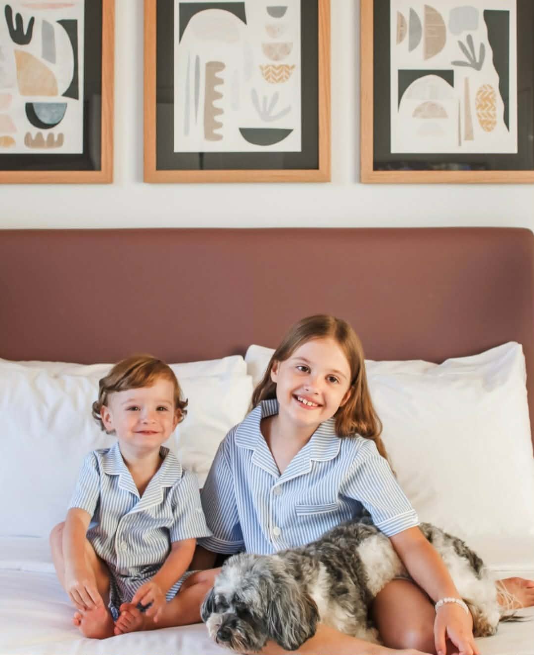 Two children in striped pajamas sit on a bed with a fluffy gray dog, smiling in a bright bedroom with abstract framed art above the headboard.