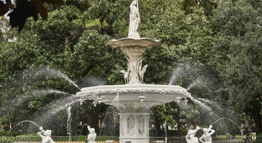 White marble, multi-tier fountain with statue on top, water sprays from the basin, surrounded by green trees and a decorative iron fence in the foreground.