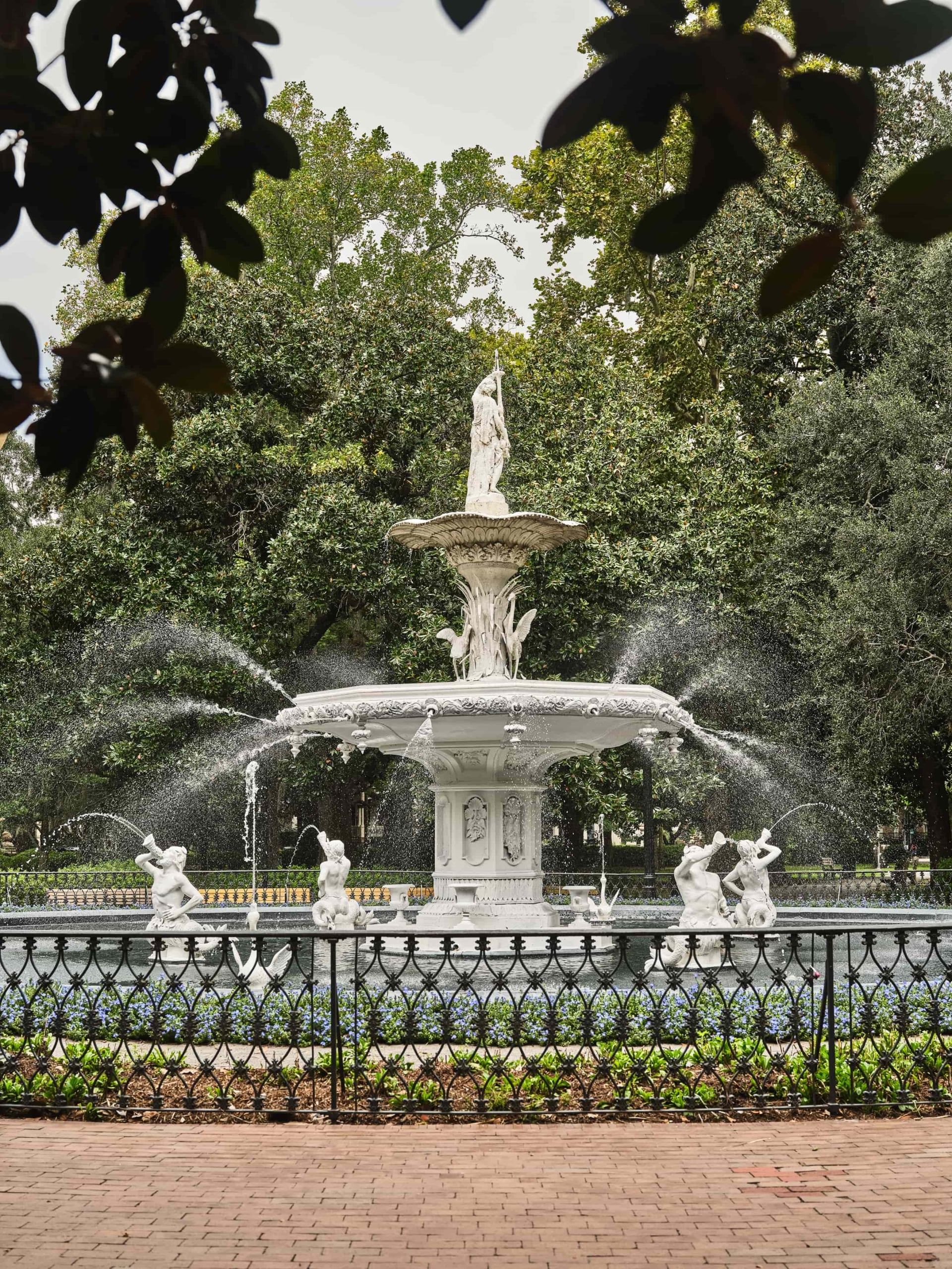 White marble, multi-tier fountain with statue on top, water sprays from the basin, surrounded by green trees and a decorative iron fence in the foreground.