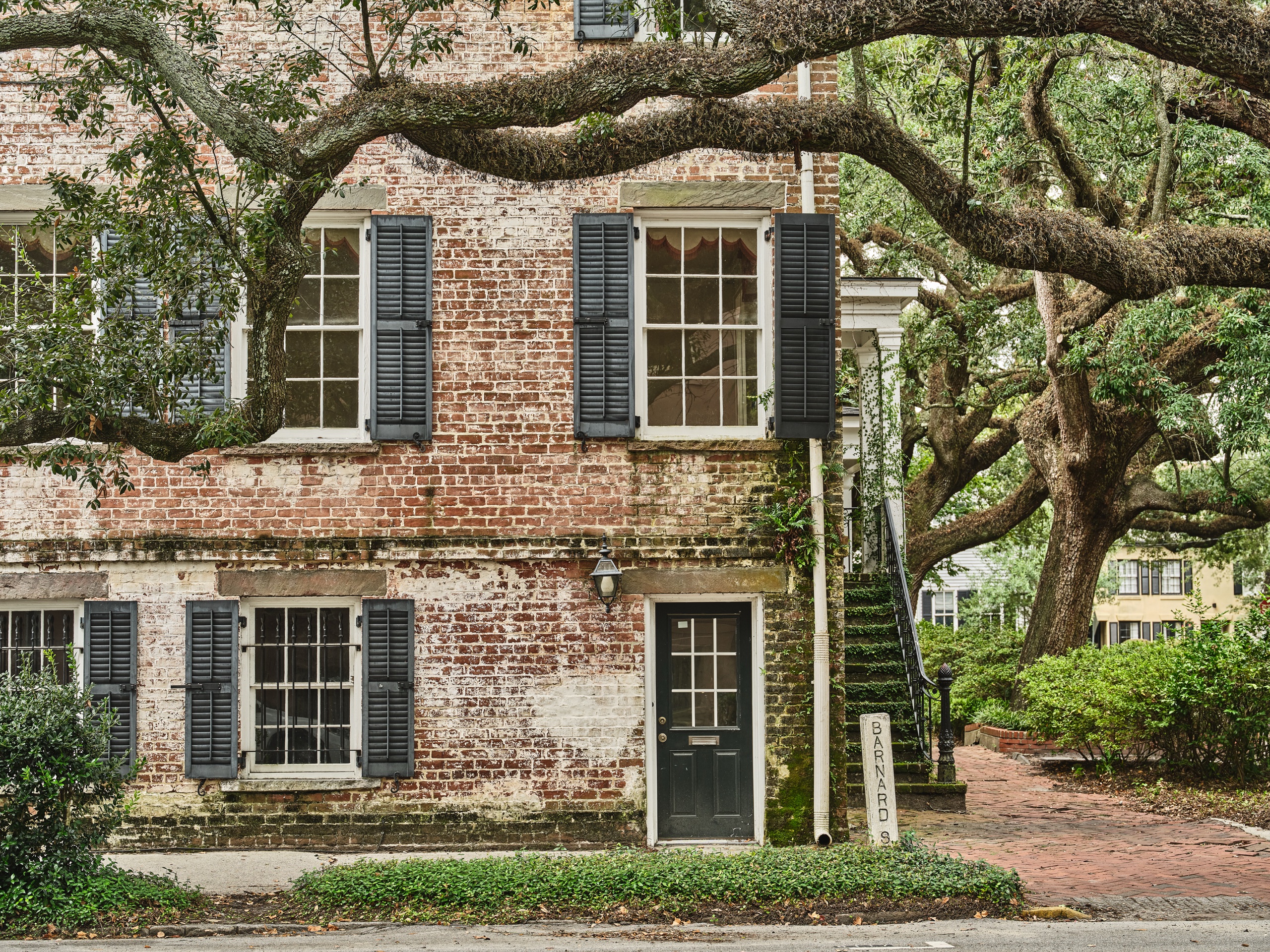 Weathered two-story brick building with dark shutters on Barnard Street, framed by a sweeping live oak tree in Savannah, GA.