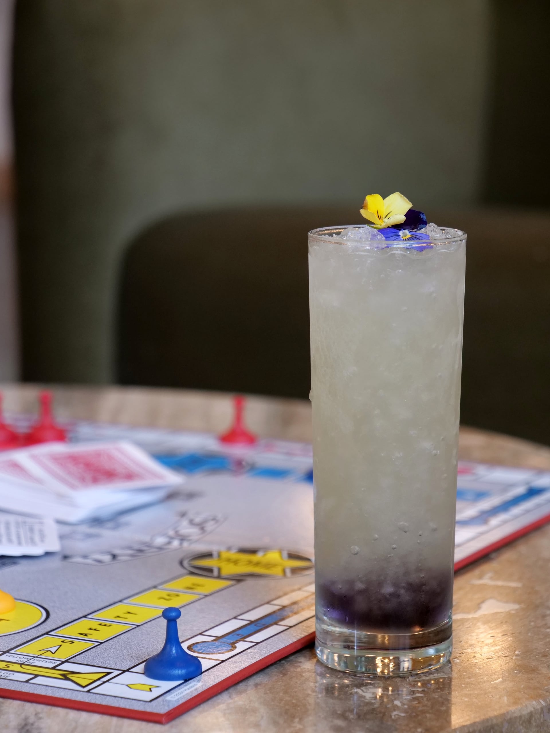 Tall frosted glass filled with crushed ice and a yellow-purple flower garnish on a colorful game-board surface (Monopoly-like).