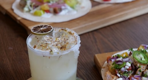 A lime margarita with a salted rim and lime slice in the foreground, tacos blurred in the background on a wooden board.