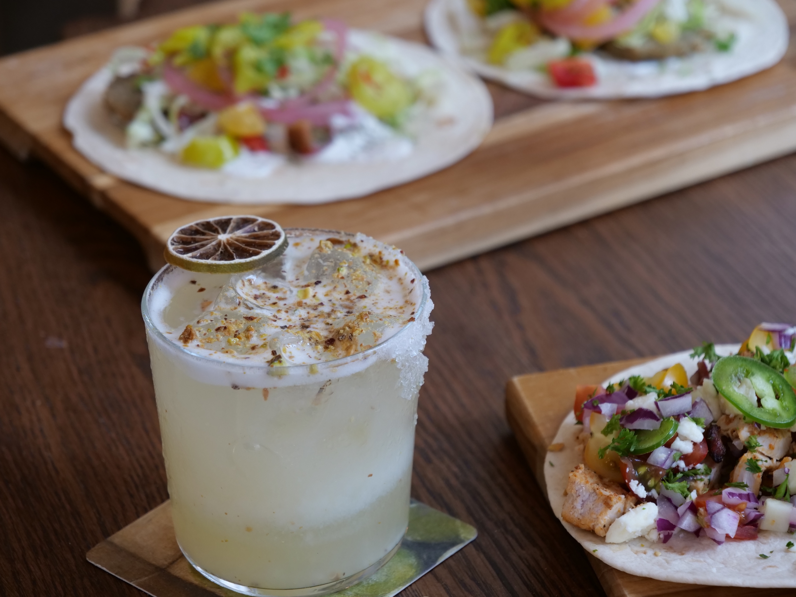 A lime margarita with a salted rim and lime slice in the foreground, tacos blurred in the background on a wooden board.