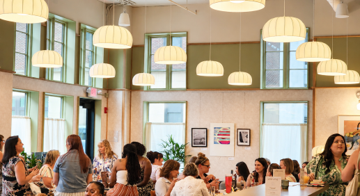 People socialize around tables in a bright cafe-like room with large hanging lamps and green-framed windows behind them.