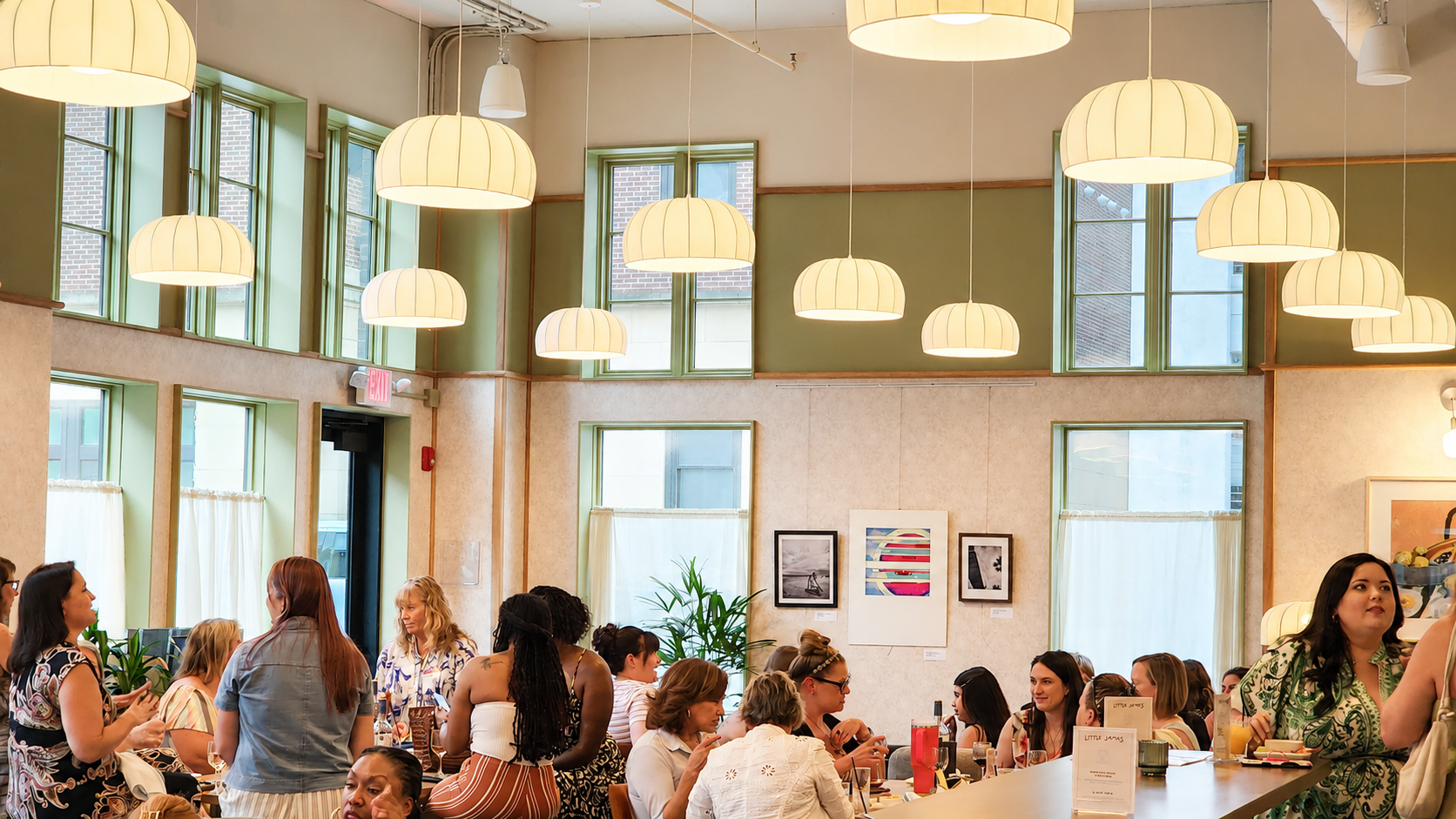 People socialize around tables in a bright cafe-like room with large hanging lamps and green-framed windows behind them.