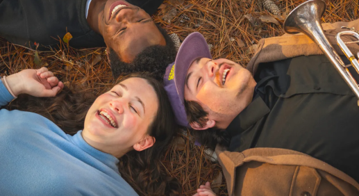 Three friends lying on pine needles in a circle, smiling and laughing outdoors in autumn.