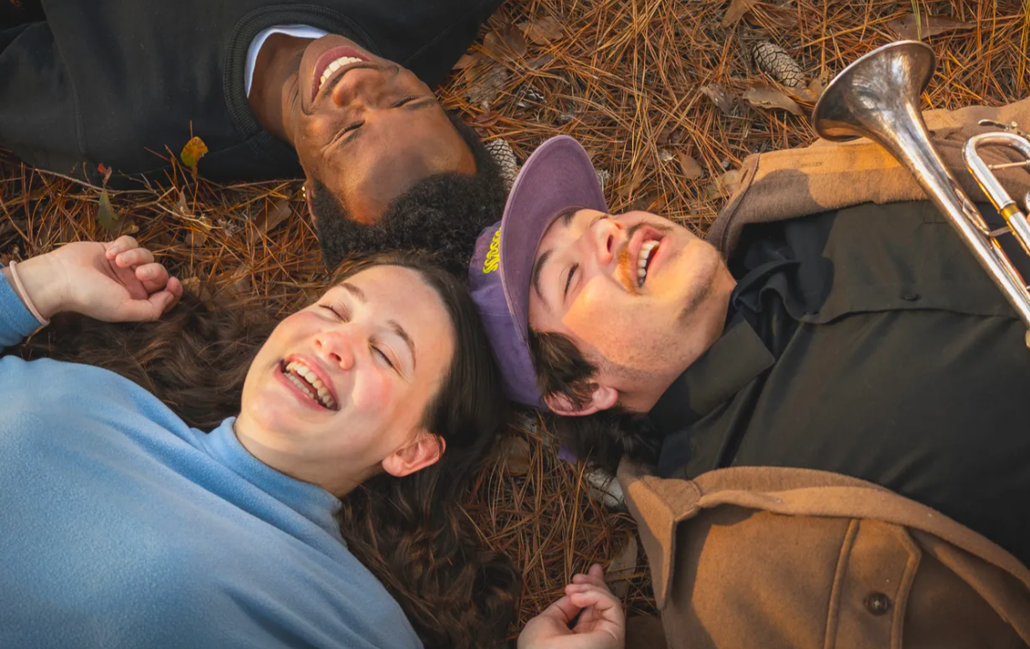 Three friends lying on pine needles in a circle, smiling and laughing outdoors in autumn.
