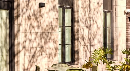 Olive-green bistro table and chairs on a sunlit courtyard patio beside a limestone building with dappled tree shadows.