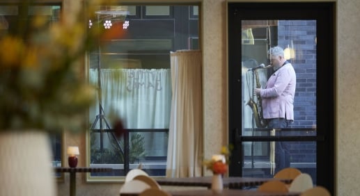 Saxophonist in a pink blazer performing outside The Ann's glass-fronted dining room during its grand opening, with candlelit tables and floral arrangements visible inside