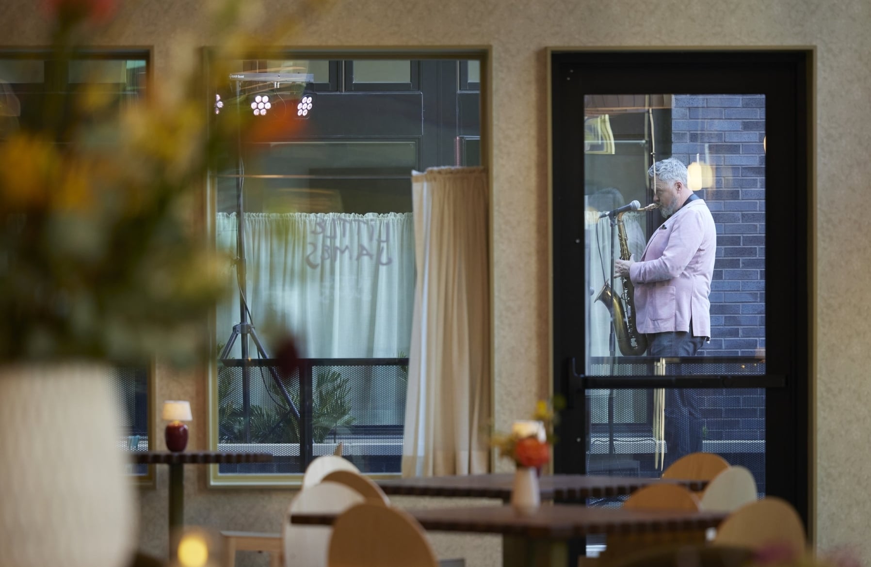 Saxophonist in a pink blazer performing outside The Ann's glass-fronted dining room during its grand opening, with candlelit tables and floral arrangements visible inside