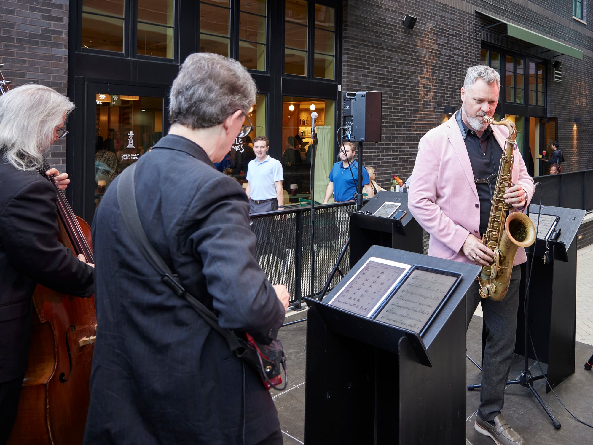 Live jazz trio performing outdoors at The Ann's grand opening celebration, with a saxophonist in a pink blazer, a bassist, and a musician reading sheet music