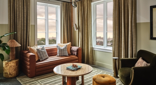 Cozy living room with a brown leather sofa, striped cushions, and a round wooden coffee table by large windows with beige curtains.