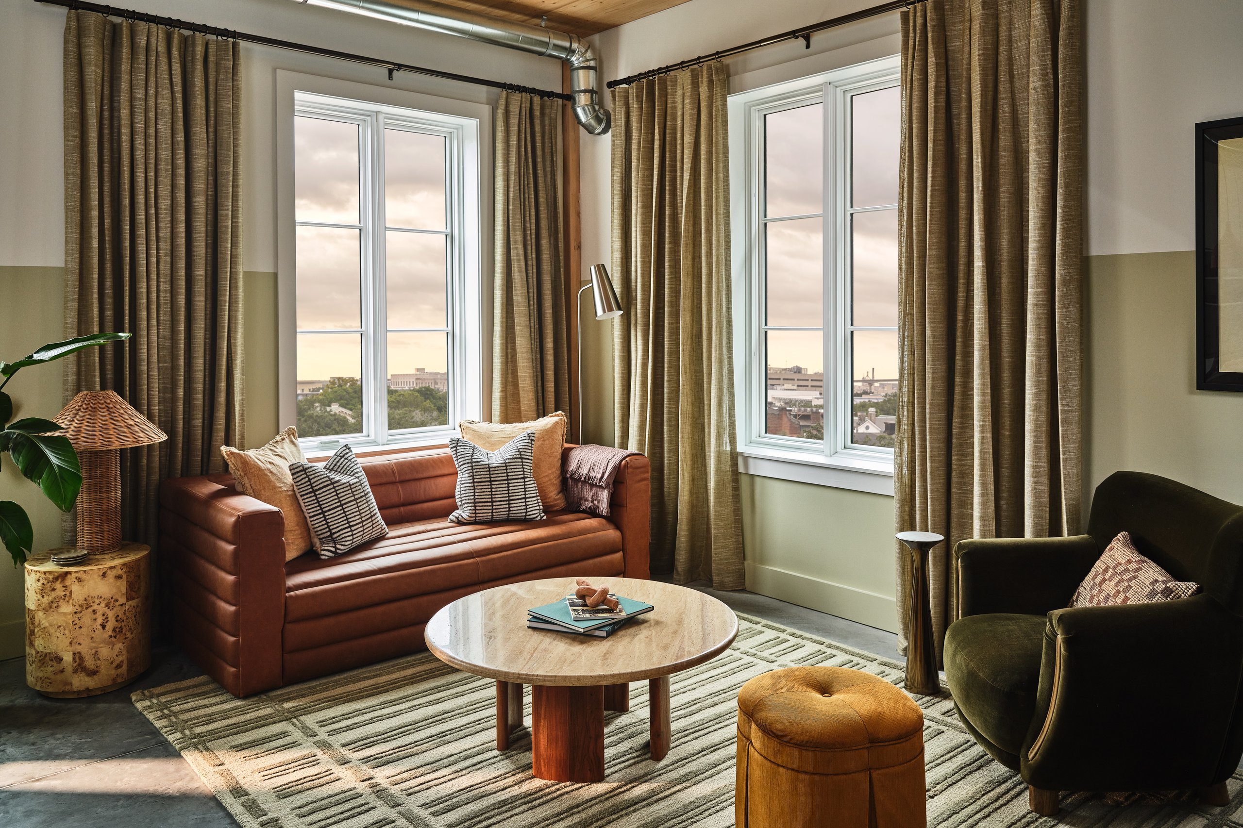 Cozy living room with a brown leather sofa, striped cushions, and a round wooden coffee table by large windows with beige curtains.