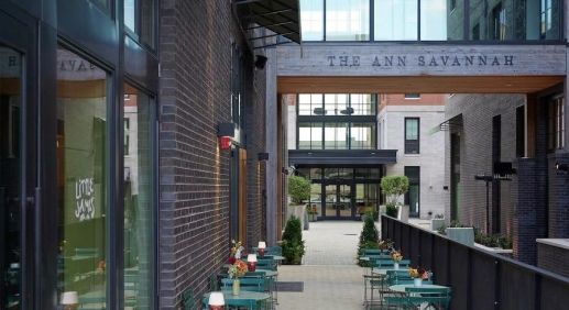 Herringbone-paved passage at The Ann Savannah with teal bistro tables, red lamps and a wood sky bridge overhead