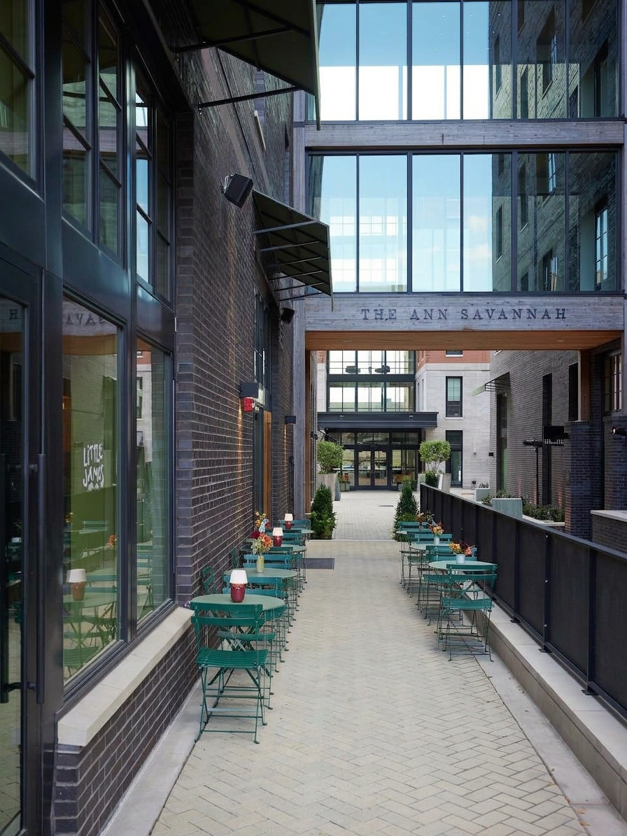 Herringbone-paved passage at The Ann Savannah with teal bistro tables, red lamps and a wood sky bridge overhead